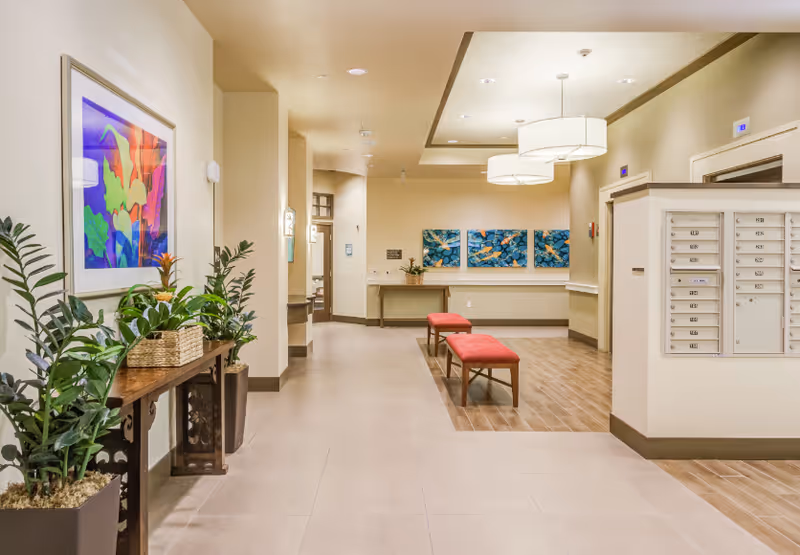 Interior hallway of The Plaza at Pearl City featuring a row of mailboxes on the right, two red cushioned benches in the center, potted plants on a wooden table to the left, colorful artwork on the walls, and ceiling lights illuminating the space.