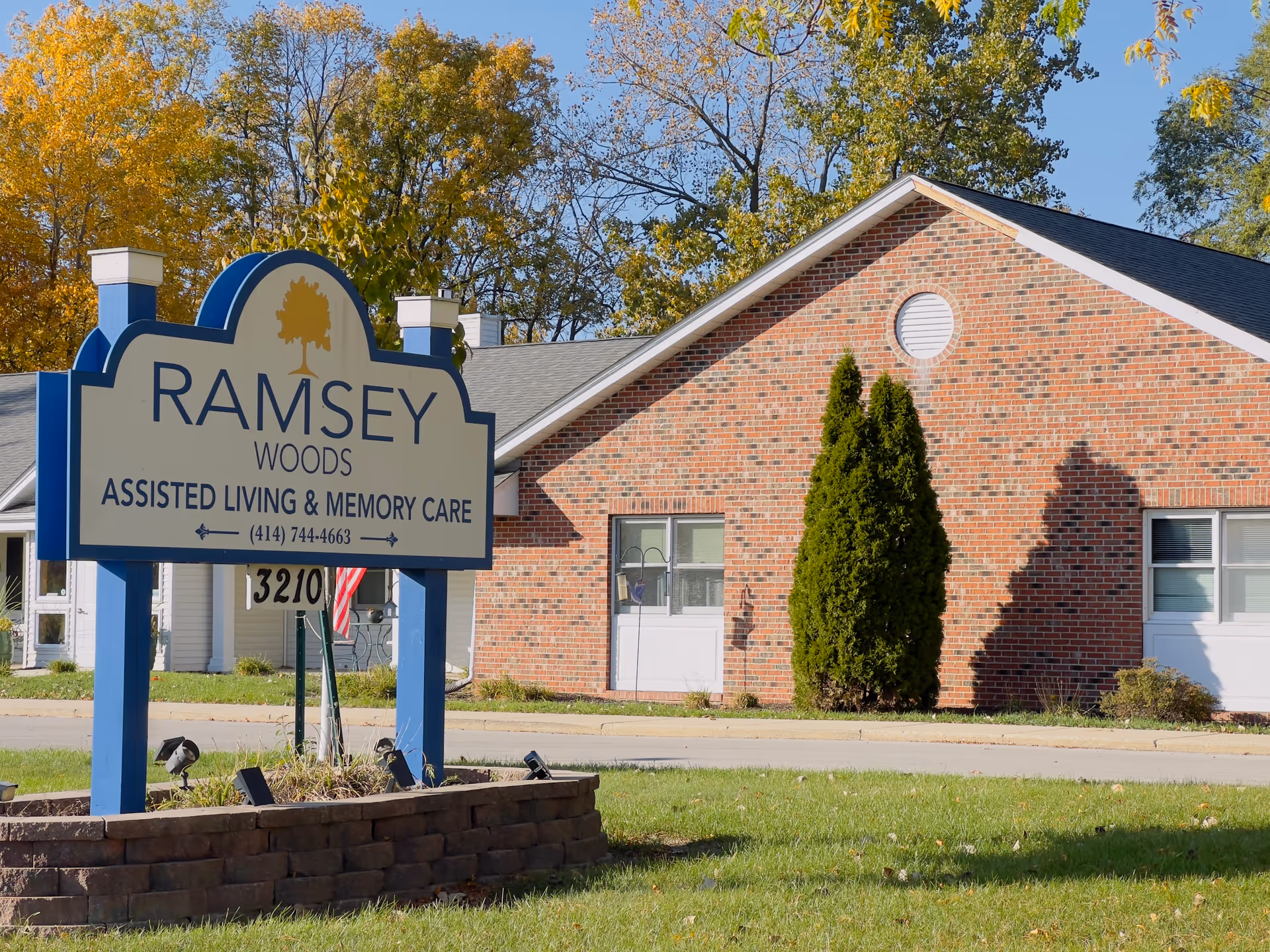 Outdoor view of Ramsey Woods assisted living and memory care facility sign with a brick building and trees in the background under a clear blue sky.