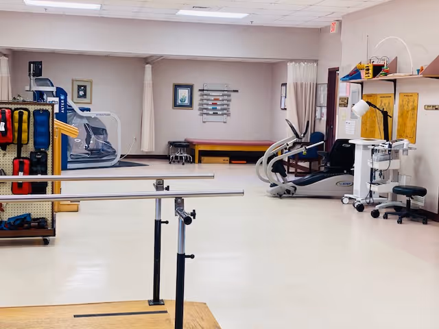 Physical therapy room with parallel bars in the foreground, exercise equipment including a treadmill and stationary bike, a treatment table, and various therapy tools and charts on the walls.