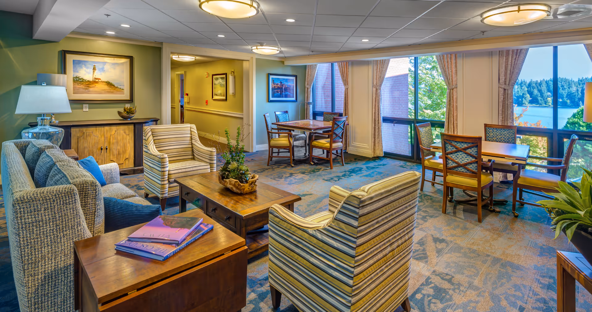 A bright and inviting common area in a senior living facility featuring comfortable seating including a sofa and striped armchairs around a wooden coffee table with a plant centerpiece. There are also two wooden tables with chairs near large windows that offer a scenic view of trees and a body of water outside. The room is decorated with framed artwork, lamps, and has carpeted flooring.