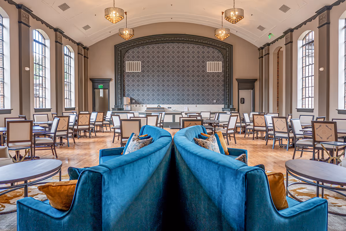 Spacious senior living dining room with large arched windows, multiple tables and chairs, and two blue velvet sofas with decorative pillows in the center. The room features a high vaulted ceiling with hanging chandeliers and a patterned accent wall at the far end.