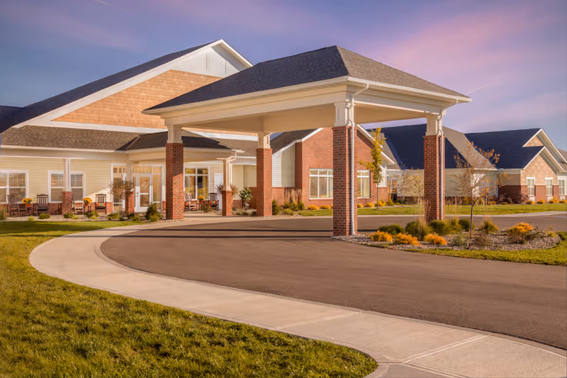 Exterior view of Randall Residence of West Milton showing a covered driveway entrance with brick pillars, surrounded by landscaped grass and shrubs, under a clear sky during sunset.