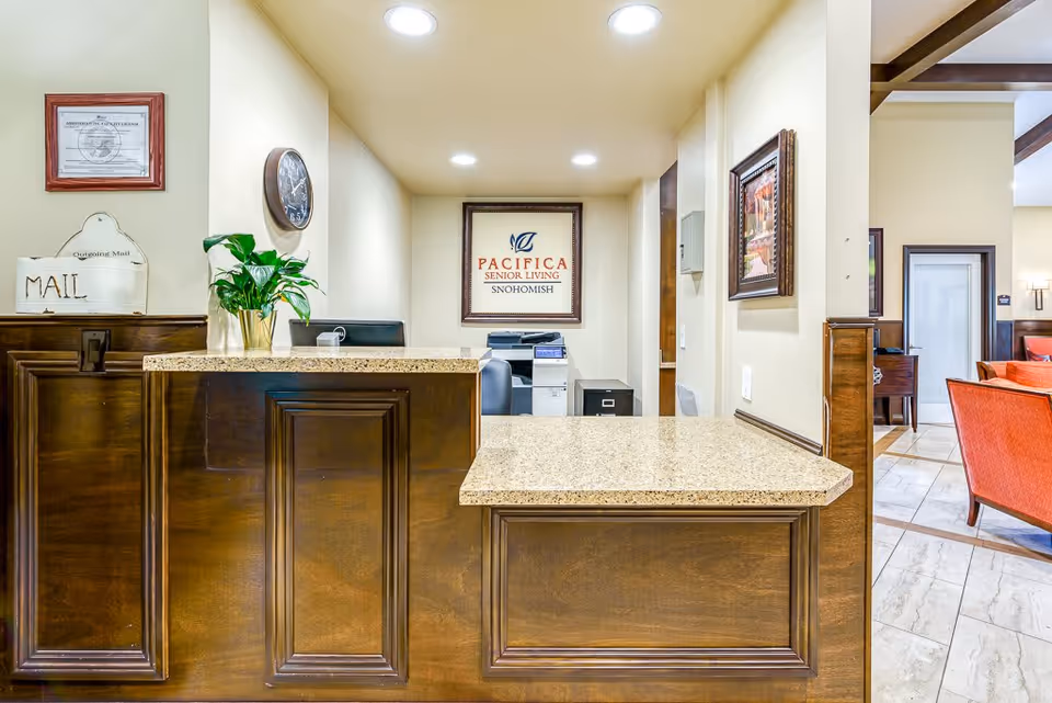 Reception desk area with a granite countertop and dark wood paneling. Behind the desk is a framed sign that reads 'Pacifica Senior Living Snohomish'. A clock is mounted on the wall to the left, along with a small plant and a mail organizer labeled 'MAIL'. To the right, there is a glimpse of a seating area with orange chairs and a piano in the background.