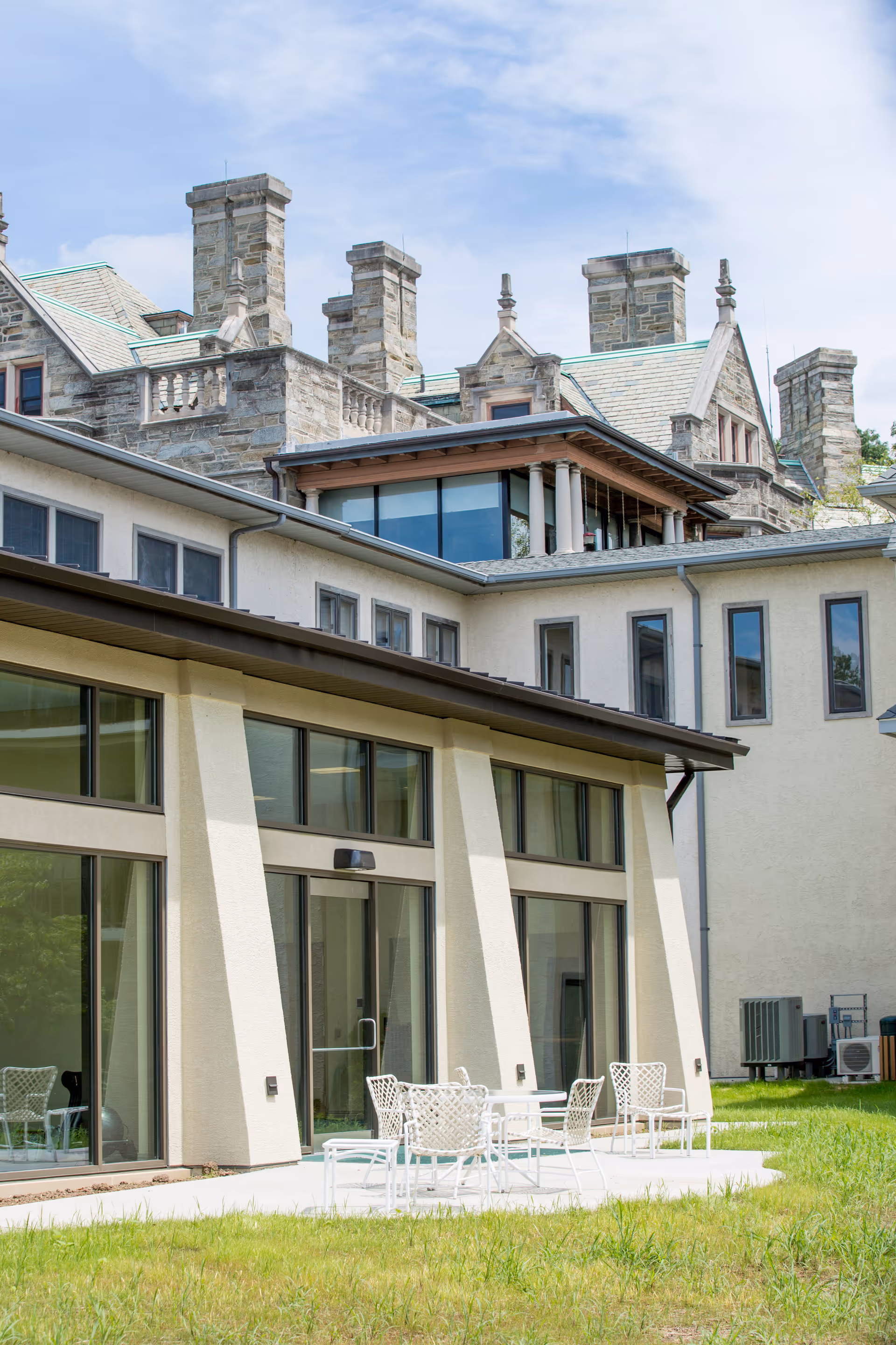 Outdoor patio area with white metal tables and chairs on a concrete surface next to a building with large windows. The building has a modern design with beige walls and angled supports. In the background, there is an older stone building with multiple chimneys and a greenish roof under a partly cloudy sky.