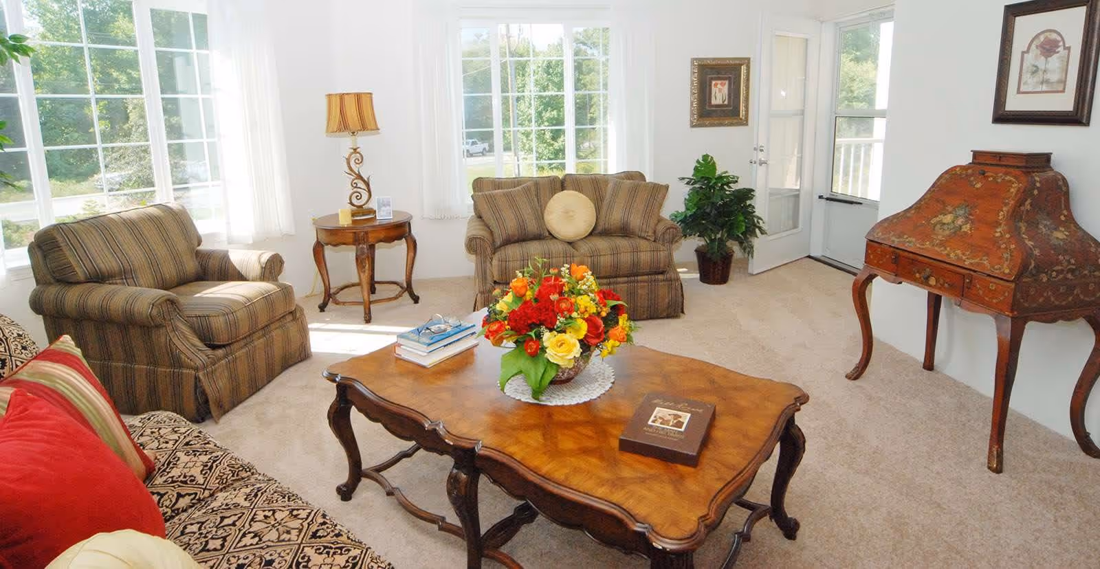 A bright and cozy living room with large windows letting in natural light. The room features a patterned sofa with red and beige cushions, two striped armchairs, a wooden coffee table with a floral centerpiece and books, a side table with a decorative lamp, a potted plant near a glass door, and a vintage wooden writing desk against the wall. The walls are white and decorated with framed artwork.