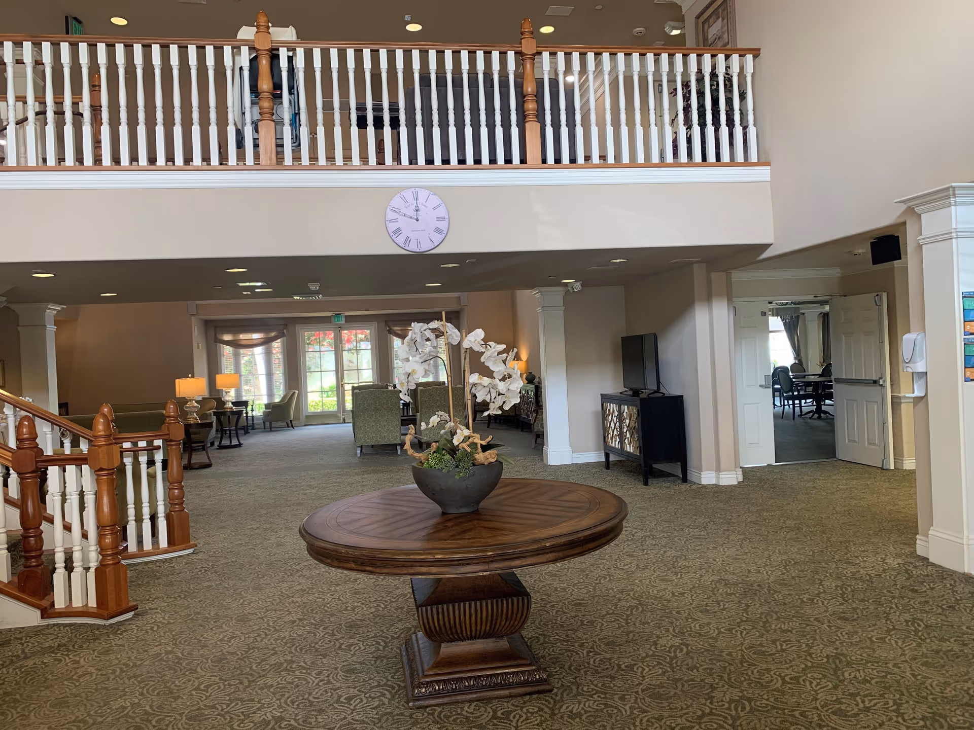 Spacious senior living facility common area with a round wooden table holding a flower arrangement in the center. The room features carpeted floors, a staircase with wooden banisters, a balcony railing above, a clock on the wall, several armchairs and tables with lamps near large windows letting in natural light, and a TV on a stand near an open doorway leading to another room.