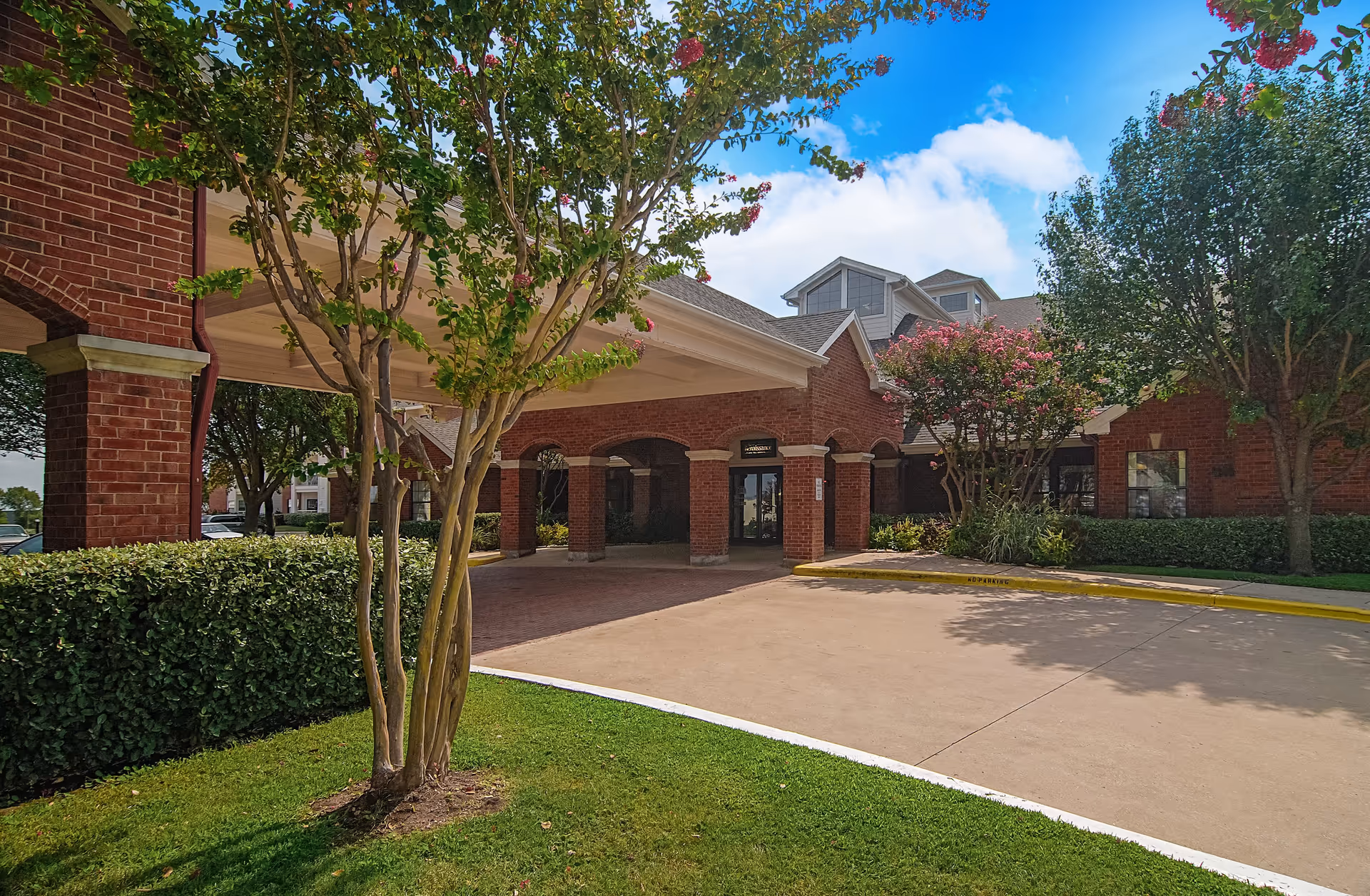 Brick building front with a covered drive-up entrance, landscaped trees and a paved driveway under a blue sky.