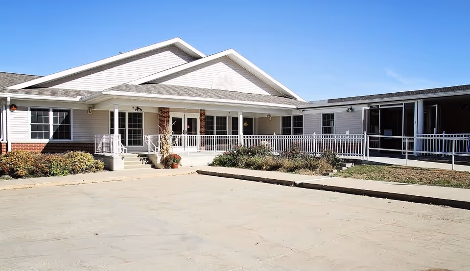 Exterior view of Griswold Rehabilitation & Health Care Center showing a single-story building with white siding and brick accents, a covered entrance with white railings, steps, and a wheelchair ramp, surrounded by some shrubs and plants under a clear blue sky.