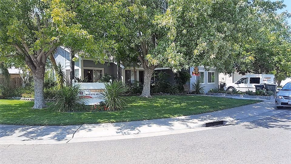 Front exterior view of Siebenthal Elder Care Home showing a residential-style building with a porch, surrounded by green trees and a well-maintained lawn. A sign with the facility's name and phone number is visible near the sidewalk. Several vehicles are parked along the street and driveway.