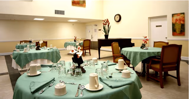 A dining room with round tables covered in green tablecloths, each set with cups, glasses, utensils, and napkins. There are wooden chairs around the tables, a piano against the wall, a clock above it, and floral paintings on the walls.