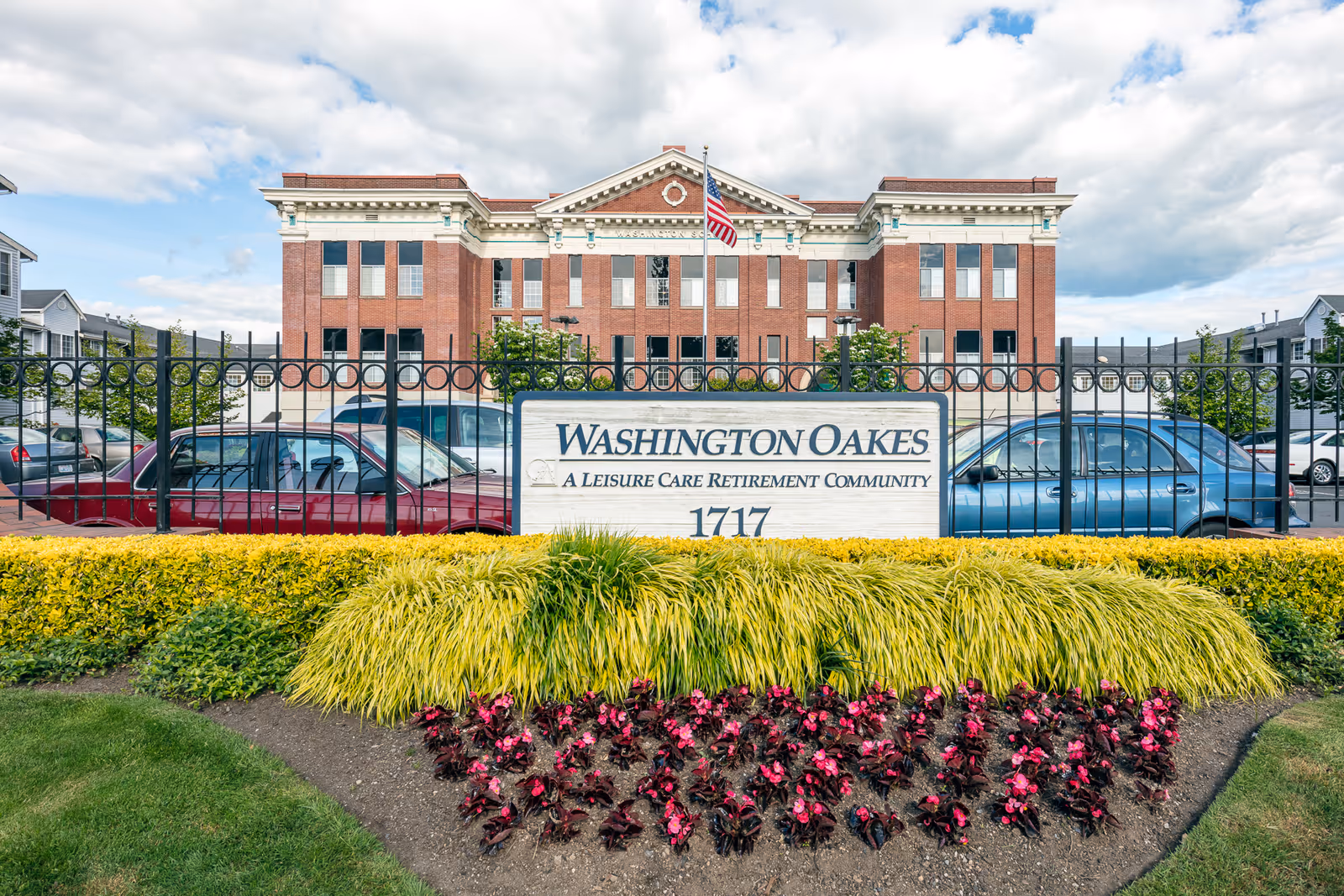 Front view of Washington Oakes, a leisure care retirement community, showing a large brick building behind a black metal fence with an American flag on a pole. In front of the fence is a landscaped area with green and yellow bushes and red flowers. A sign with the community name and address 1717 is prominently displayed.