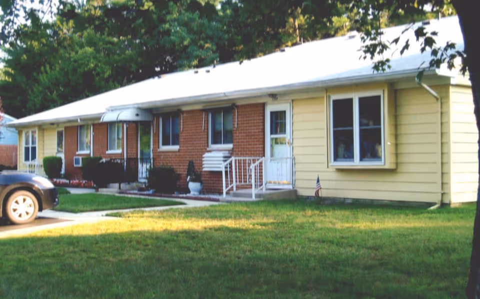 Front exterior of a single-story senior living facility with brick and beige siding, small entry steps, and a grassy lawn.