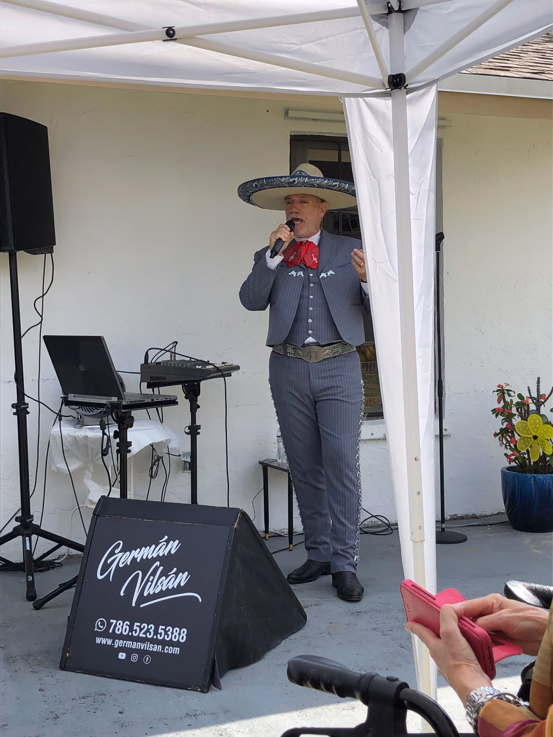 A man dressed in a traditional mariachi outfit with a large sombrero and red bow tie is singing into a microphone under a white canopy. There is sound equipment including a laptop and speaker nearby. A sign in front of the man reads 'Germán Vilsán' with contact information. A person holding a pink phone is partially visible in the foreground.