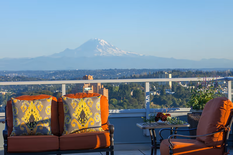 Outdoor patio seating area with orange cushioned chairs and patterned pillows, a small table with a floral arrangement, overlooking a scenic view of a city and distant snow-capped mountain under a clear blue sky.