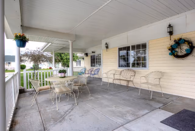 Covered outdoor patio area with metal chairs and tables, hanging flower pots, a wreath on the wall, and a view of a grassy lawn and neighboring buildings.
