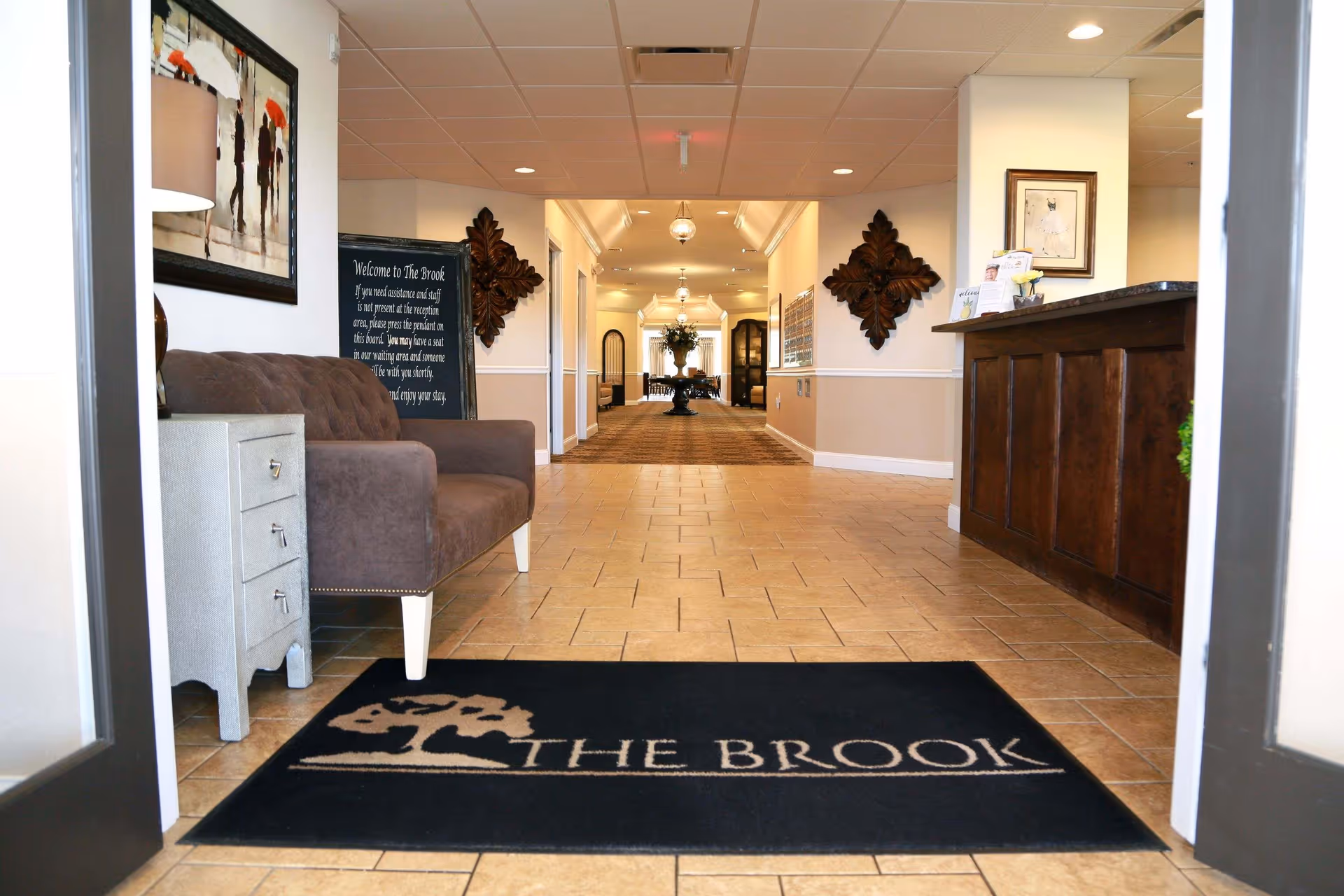Entrance area of The Brook of Big Rapids featuring a black floor mat with the facility's logo, a brown upholstered bench next to a small gray drawer unit, a wooden reception desk on the right, and a long hallway with decorative wall art and lighting fixtures.