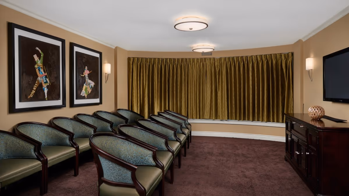 A small meeting or presentation room with two rows of green cushioned chairs with wooden frames facing a dark wooden cabinet with a flat-screen TV mounted above it. The room has beige walls, two framed artworks on the left wall, wall sconces, a brown carpet, and a large window covered by gold curtains.