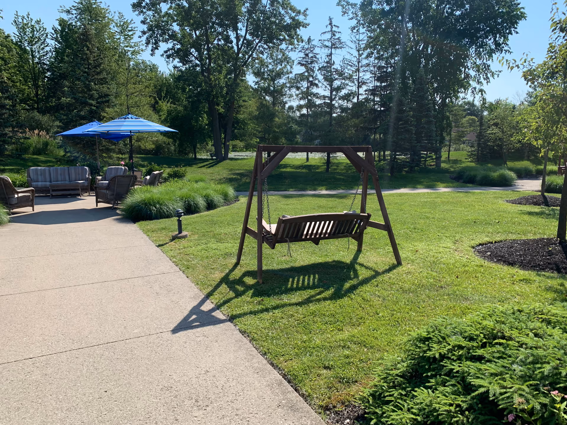 Outdoor garden area at Solstice Senior Living at East Amherst featuring a wooden swing on a grassy lawn, a concrete pathway, and a seating area with cushioned chairs and blue umbrellas surrounded by trees and greenery.