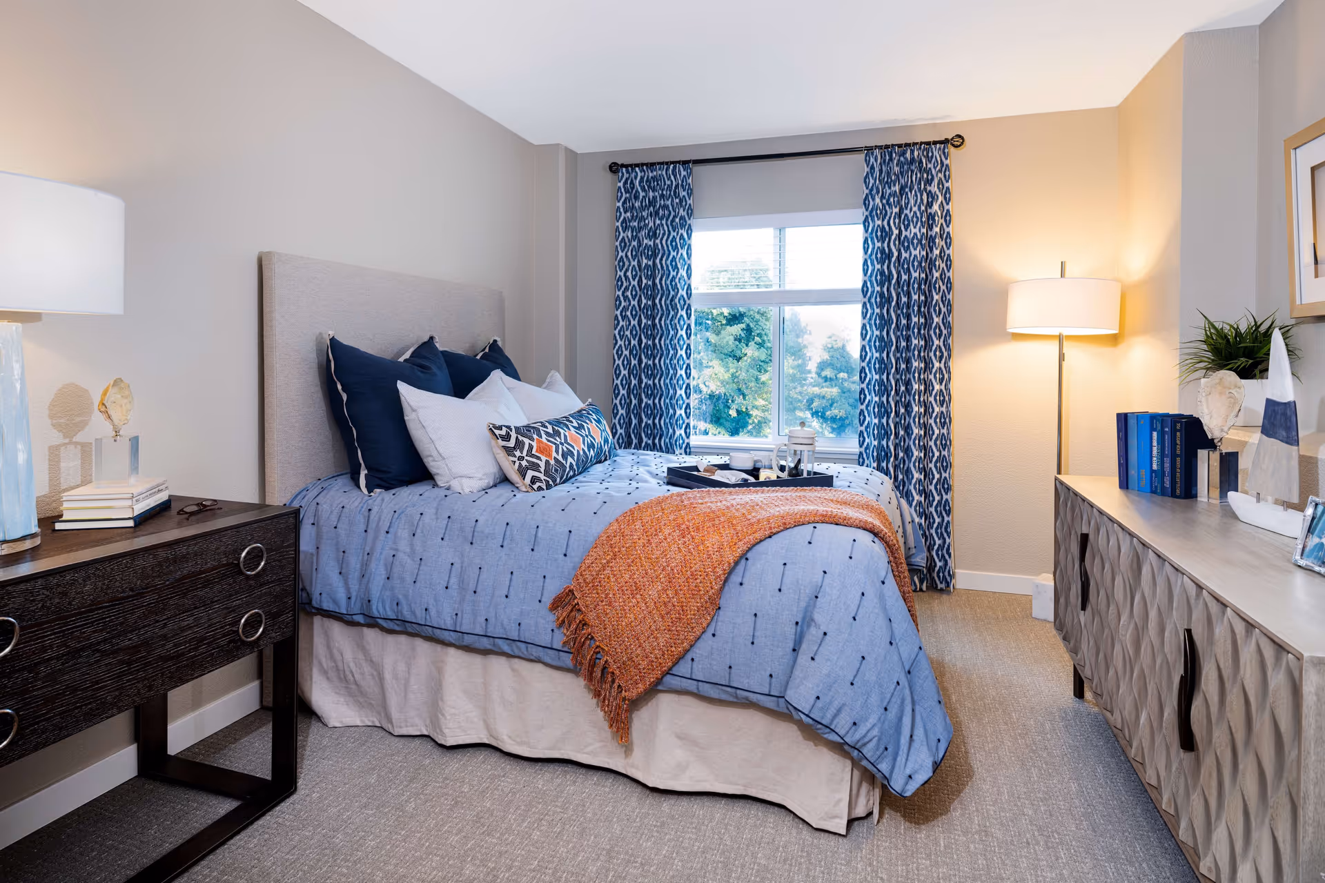 A cozy bedroom with a bed dressed in blue bedding and multiple pillows, an orange throw blanket, a window with blue patterned curtains, a dark wooden nightstand with a lamp and books, and a light-colored dresser with decorative items and a floor lamp beside it.