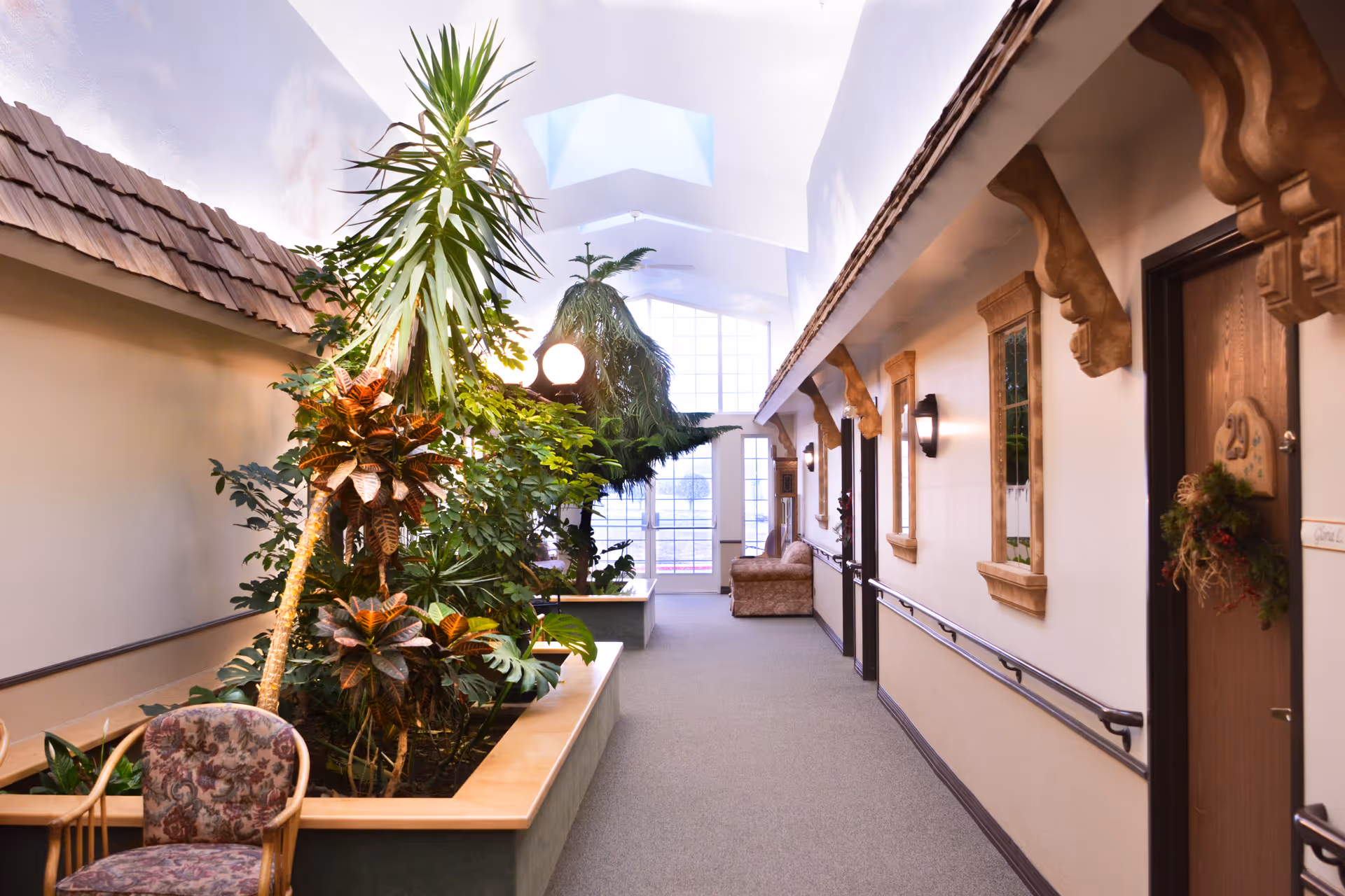 Indoor hallway of a senior living facility with a skylight ceiling, large windows at the end, and a built-in planter filled with various green plants. The hallway has handrails along the walls, wooden doors with decorative trim, and a cushioned chair and sofa placed along the corridor.