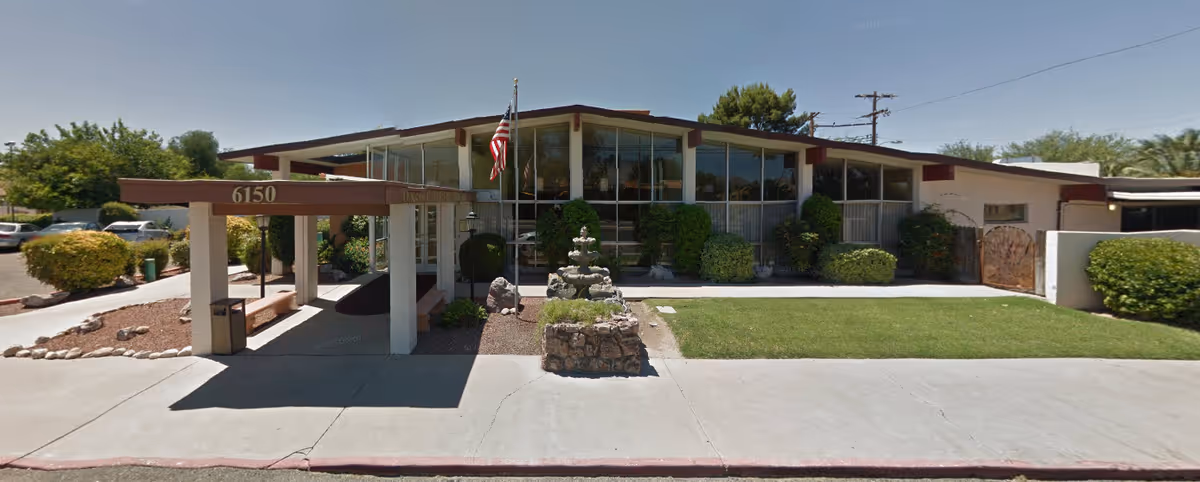 Front exterior view of Devon Gables Rehabilitation Center building with large windows, an American flag, a covered entrance with the number 6150, landscaped bushes, a small stone fountain, and a concrete sidewalk in front.