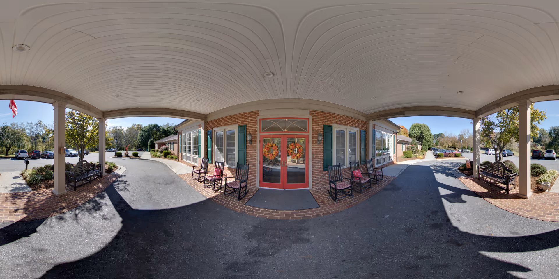Covered entrance area of a senior living facility with a red double door decorated with autumn wreaths. There are several black rocking chairs with red cushions placed along the brick wall on either side of the door. The area is paved with asphalt and brick, with benches and landscaping visible under the covered porch. Trees, parked cars, and a clear blue sky are visible in the background.