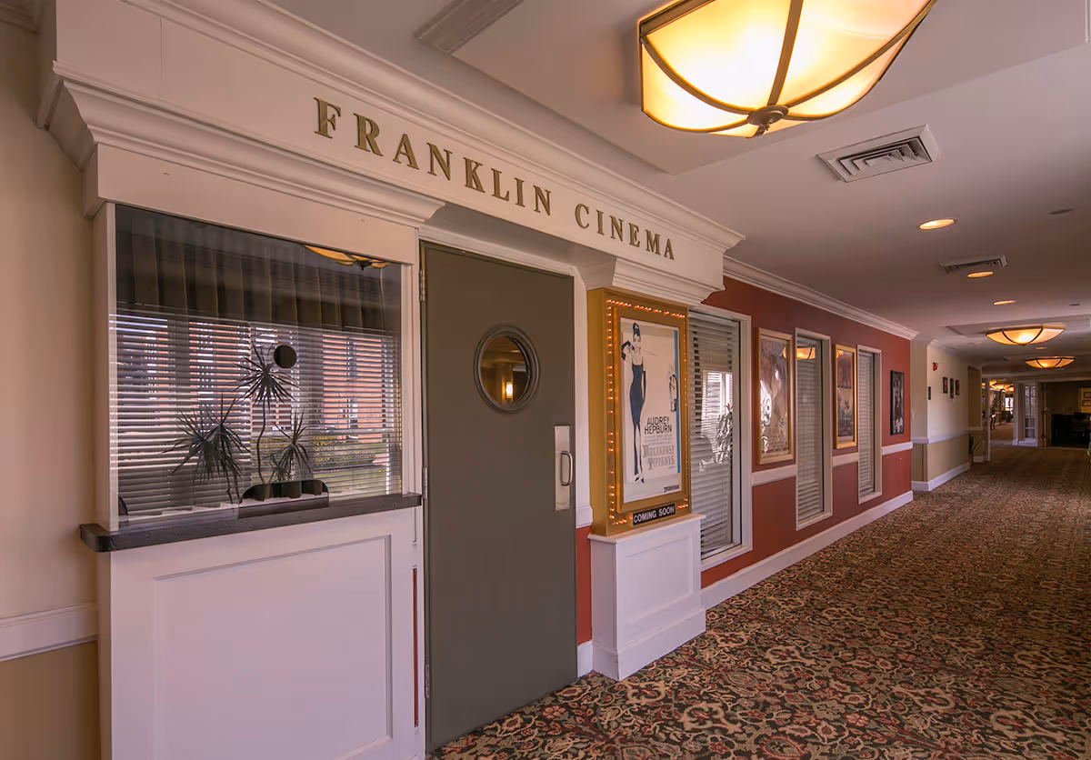 Interior hallway of a senior living facility featuring the entrance to Franklin Cinema with a ticket window, a door with a round window, and movie posters on the wall. The hallway has patterned carpet, warm lighting, and decorative molding.