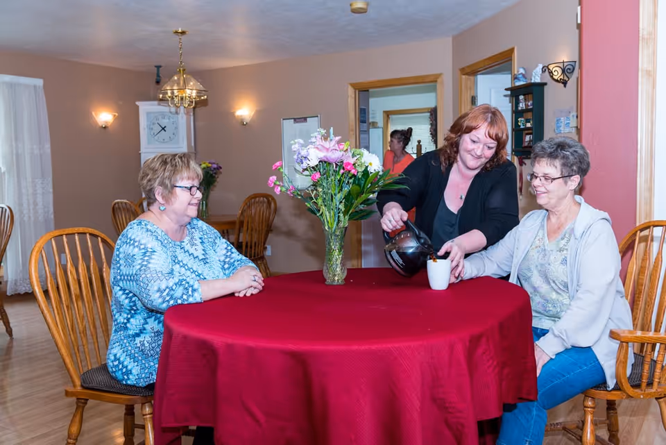 Three women sitting around a round table with a red tablecloth in a dining room. One woman is pouring coffee into a cup held by another woman, while the third woman watches and smiles. There is a vase with flowers in the center of the table. The room has wooden chairs, a clock on the wall, and warm lighting.