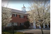 Exterior view of a two-story brick building with white trim and a cupola on the roof, surrounded by blooming trees and a paved driveway in front.