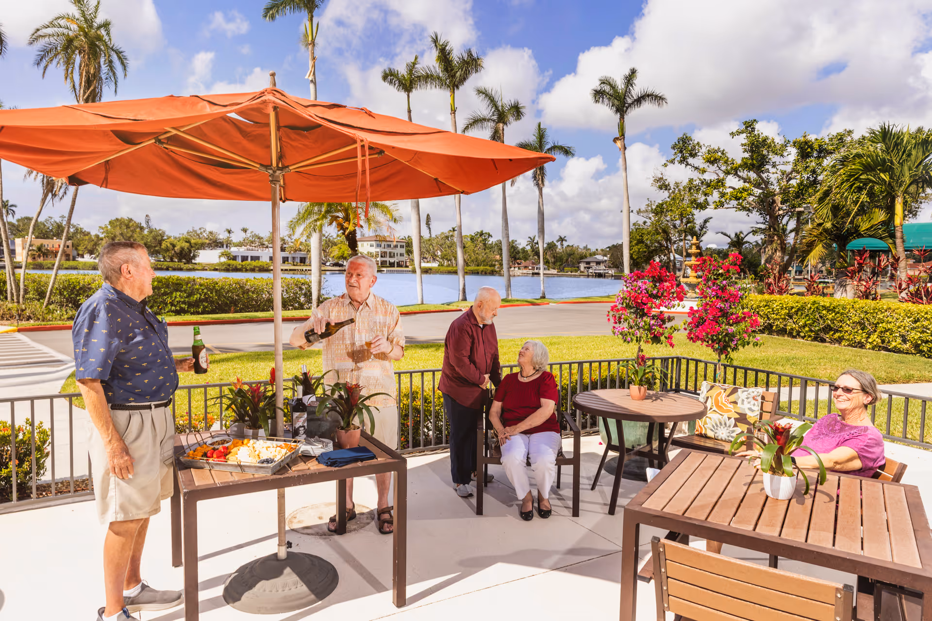 A group of elderly people socializing outdoors on a sunny day under an orange patio umbrella. Two men stand near a table with food and drinks, one pouring a beverage. Another elderly man is standing and talking to a seated woman, while another woman sits at a nearby table with a potted plant. Palm trees and a body of water are visible in the background.