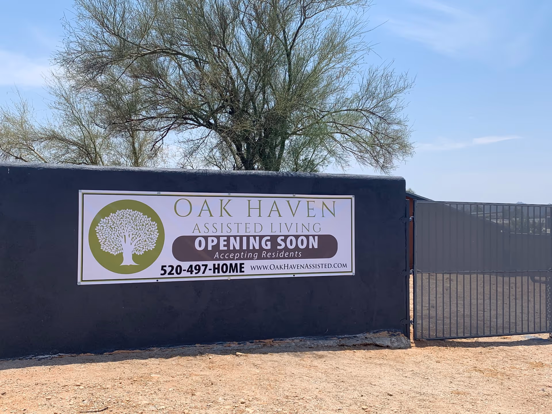 A sign on a dark wall next to a metal gate under a tree, advertising Oak Haven Assisted Living with text stating 'Opening Soon Accepting Residents' and a contact phone number and website.