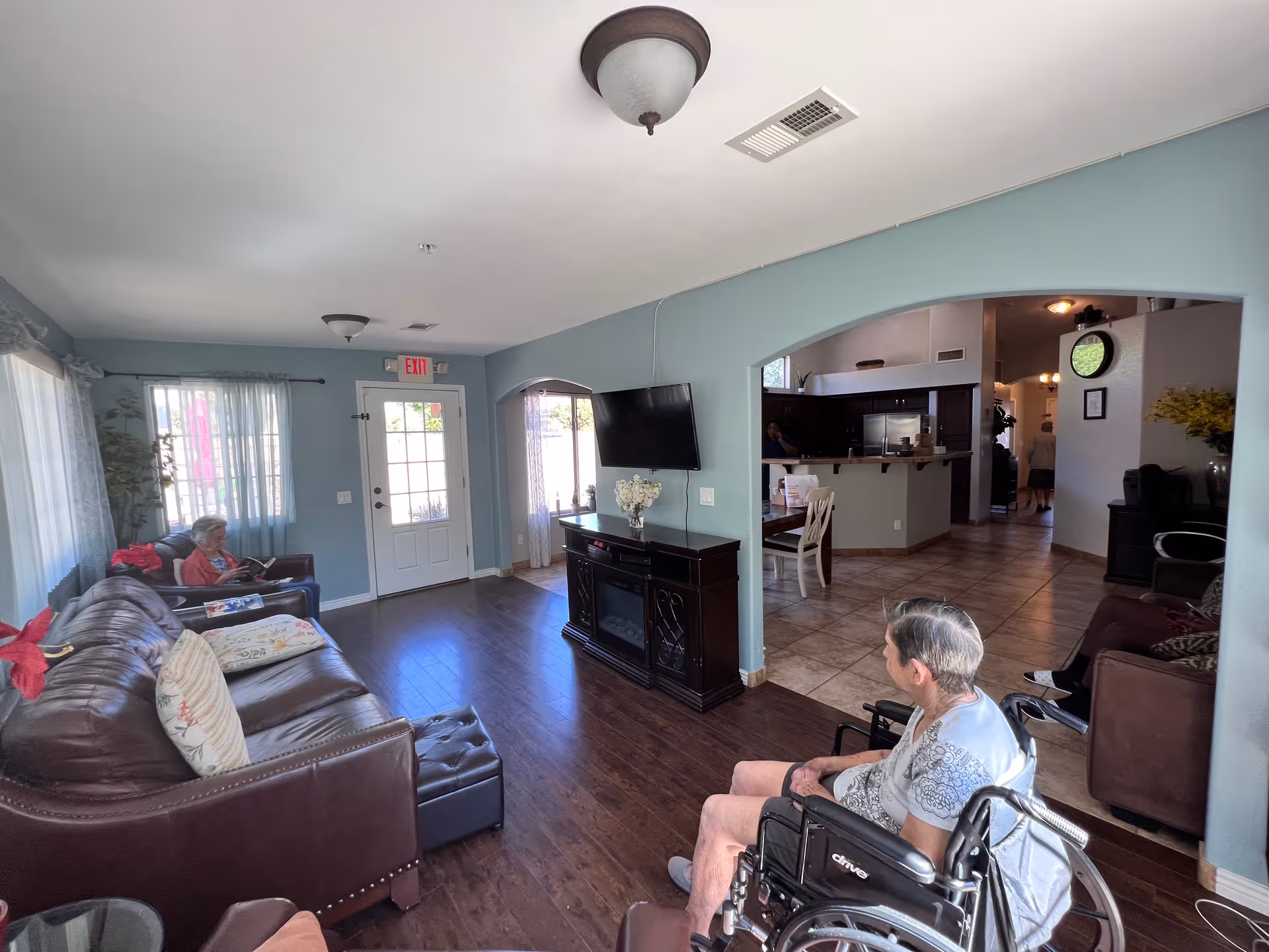 A senior living room with sofas, a wall-mounted TV, and residents seated including one in a wheelchair.