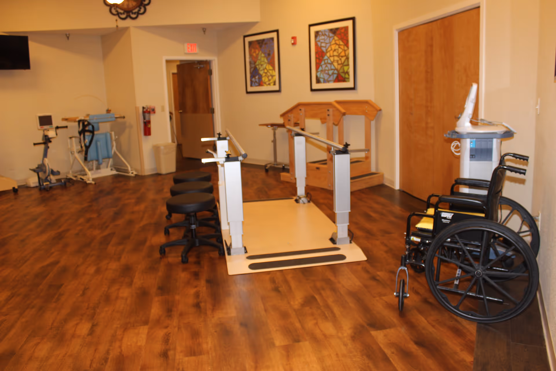 Rehabilitation room with parallel bars, wheelchairs, stools, and exercise equipment on wood flooring.
