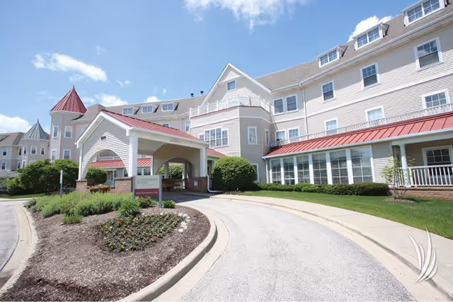 Exterior view of Victorian Village Continuing Care Senior Community building with a covered entrance, manicured landscaping, and a curved driveway under a partly cloudy sky.