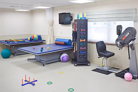 A therapy or recreational room with various exercise and activity equipment including a ping pong table, pool table, exercise balls, colorful cones, dumbbells, a seated exercise machine, and ring toss game pieces scattered on the floor. The room has light-colored walls and carpet, a wall-mounted TV, and a window with a blind.