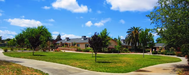 Single-story residential building set behind a grassy courtyard with a winding sidewalk, palm tree, and blue sky.