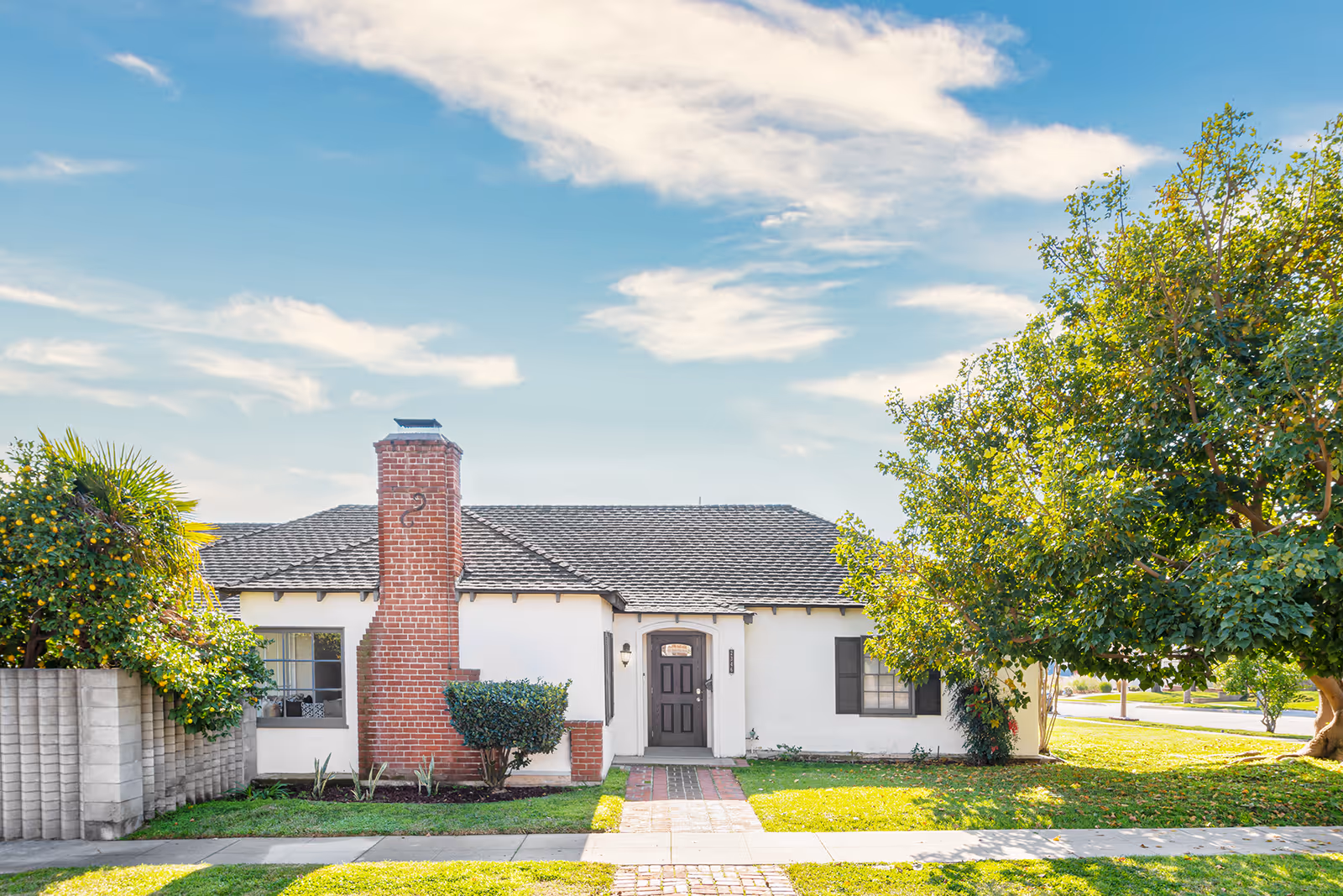 Front exterior view of a single-story house with a brick chimney, a dark front door, white walls, and a well-maintained lawn with trees and shrubs under a partly cloudy sky.