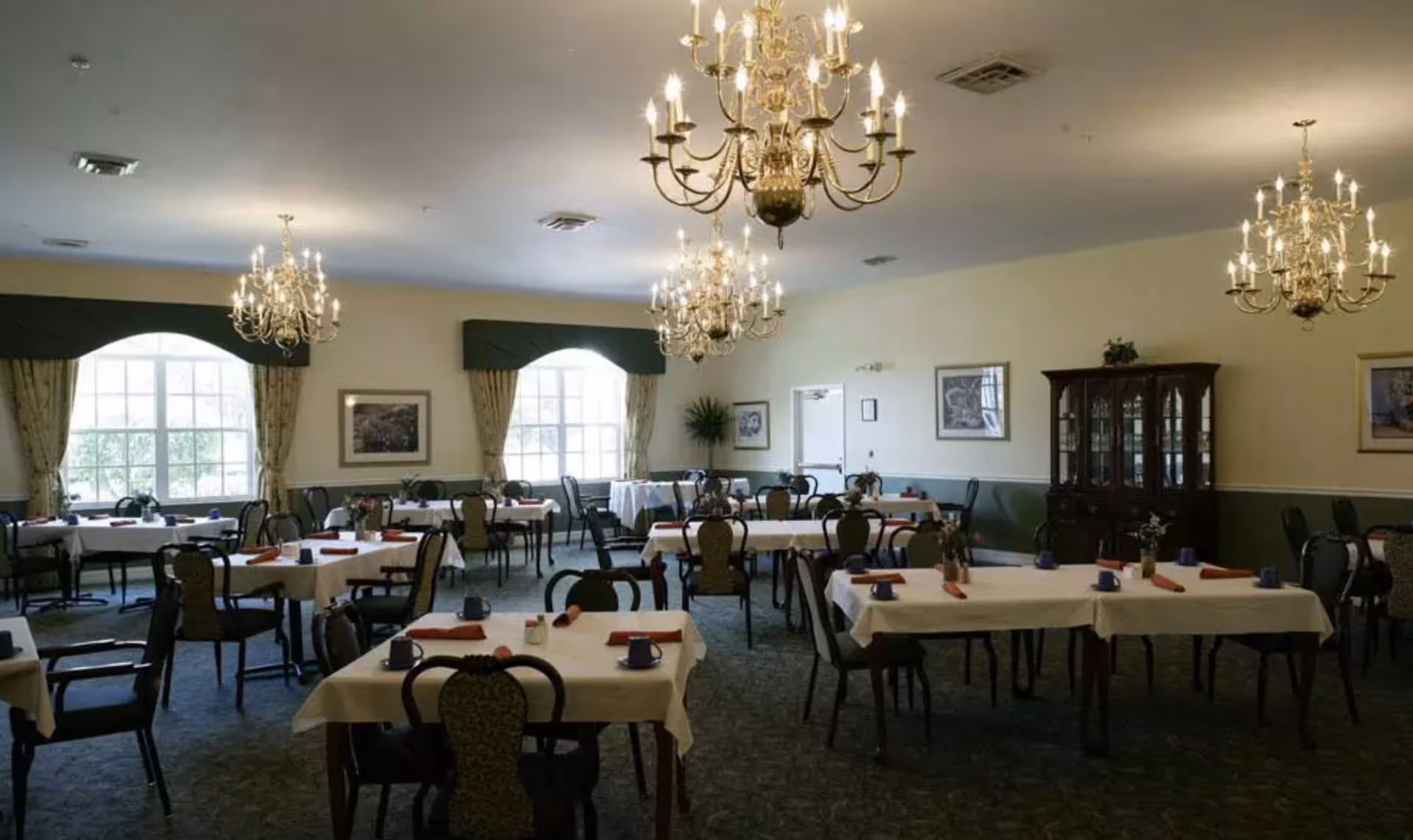 A formal dining room with multiple tables covered in white tablecloths, chairs, chandeliers, and large windows.