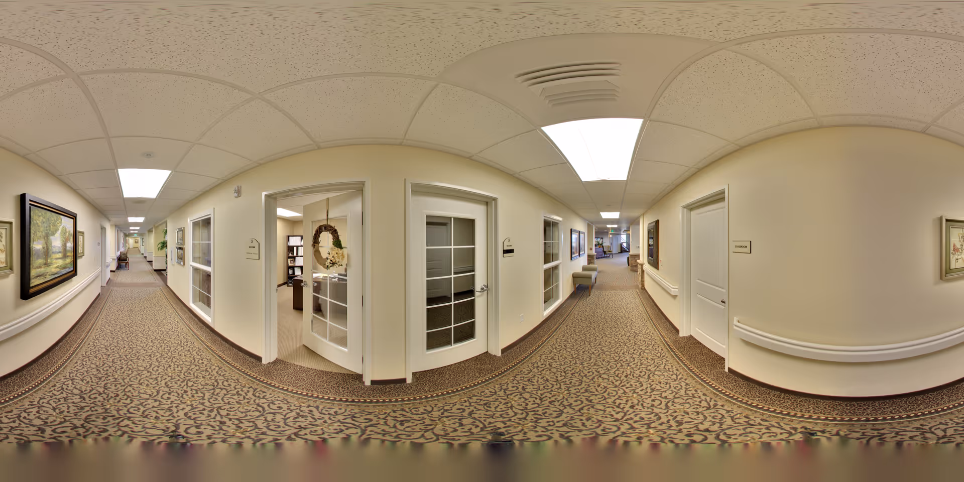 A long, carpeted hallway in a senior living facility with cream-colored walls and ceiling tiles. The hallway is decorated with framed paintings and has several doors leading to rooms, some with glass panels. Handrails run along the walls, and the lighting is bright with ceiling lights. The hallway appears clean and well-maintained.