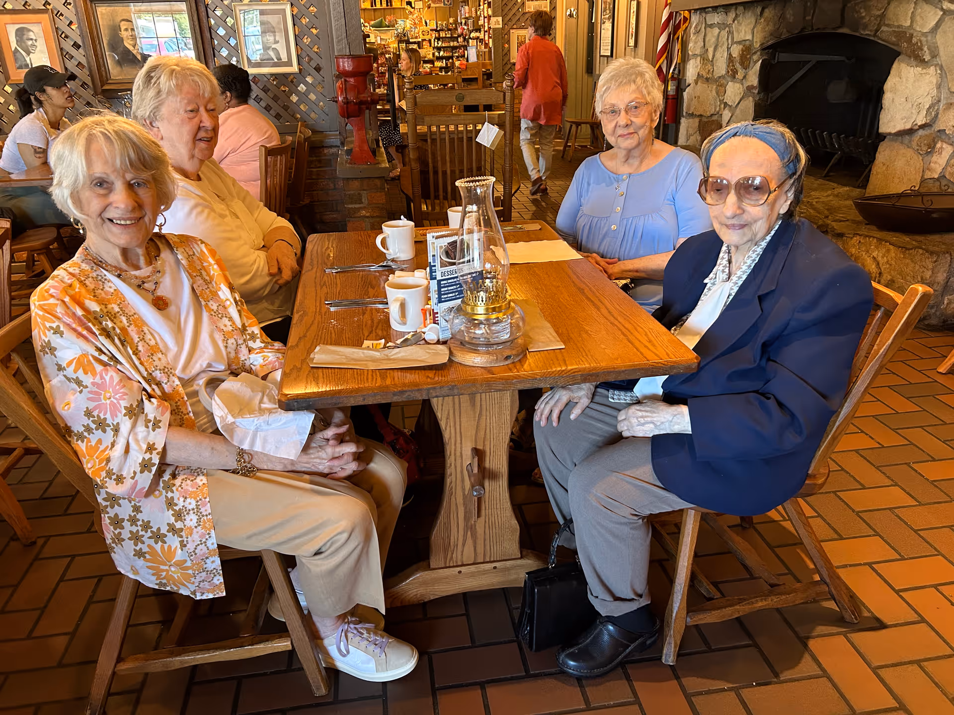 Four elderly women sitting around a wooden dining table in a cozy restaurant or dining area. The table has cups, utensils, and a glass lantern. The background shows other diners, framed pictures on the wall, and a stone fireplace.