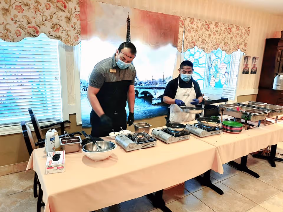 Two staff members wearing masks and gloves preparing food on portable stoves in a dining area with a table covered in a beige tablecloth. The background features a large window with a Paris-themed curtain showing the Eiffel Tower and a bridge over a river.