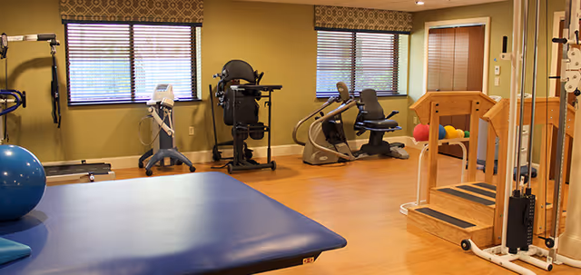 A senior rehab exercise room with a treatment table, exercise ball, recumbent bikes, strength machines and therapy stairs under windows.