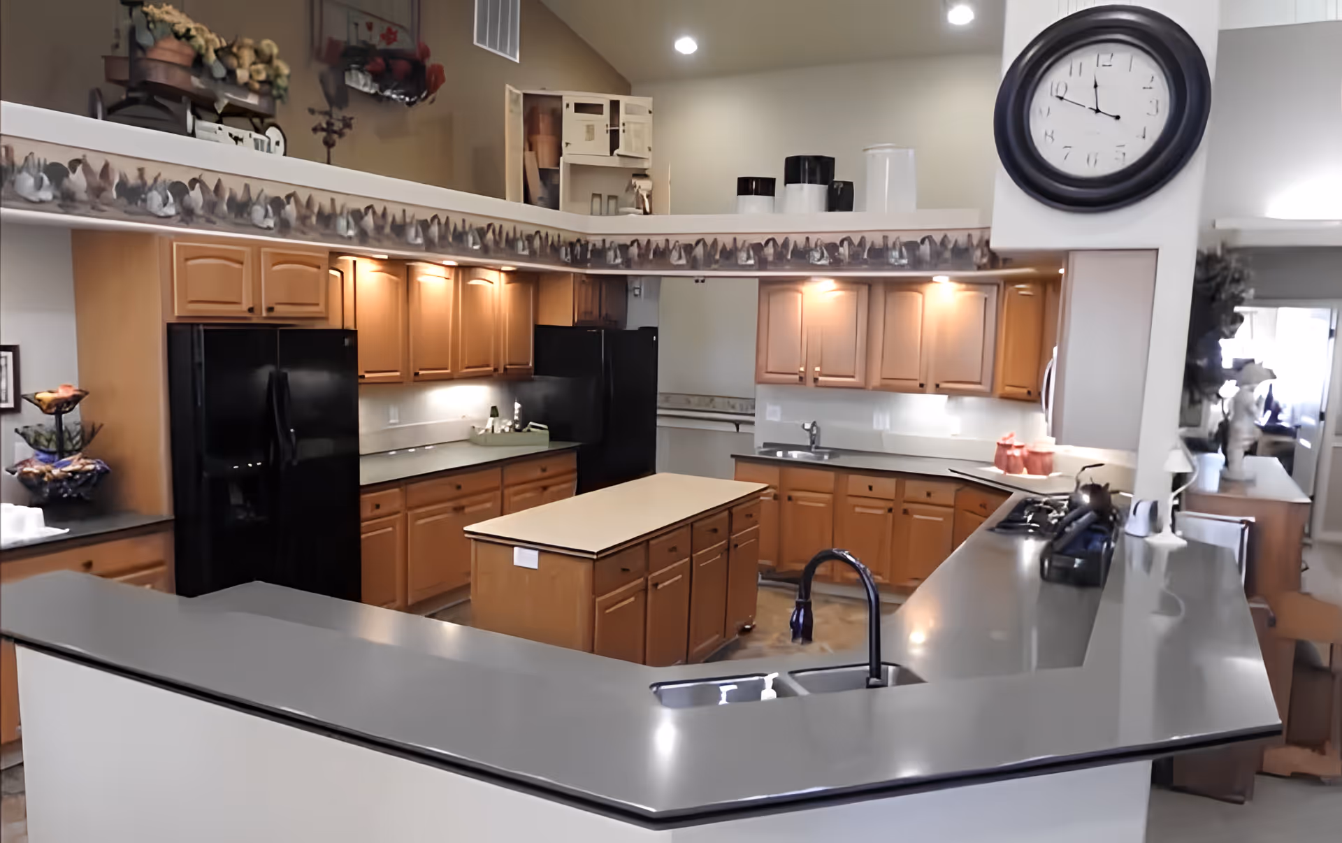 A spacious kitchen area with wooden cabinets, black appliances including two refrigerators, a central island with drawers, and a large countertop with a sink and faucet. The kitchen is well-lit with under-cabinet lighting and has a large wall clock. Decorative items and plants are placed on top of the cabinets and counters.