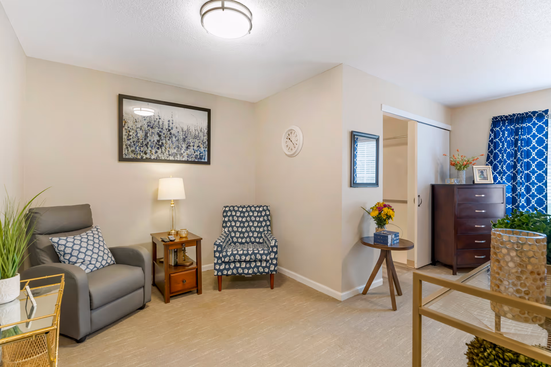A cozy living room area with a gray recliner chair and a patterned blue armchair. Between them is a wooden side table with a lamp and decorative items. On the wall above the chairs is a framed abstract artwork. To the right, there is a small round wooden table with a flower vase and a mirror hanging above it. In the background, a dark wooden dresser stands near a window with blue patterned curtains. The room has beige walls and carpeted floor.