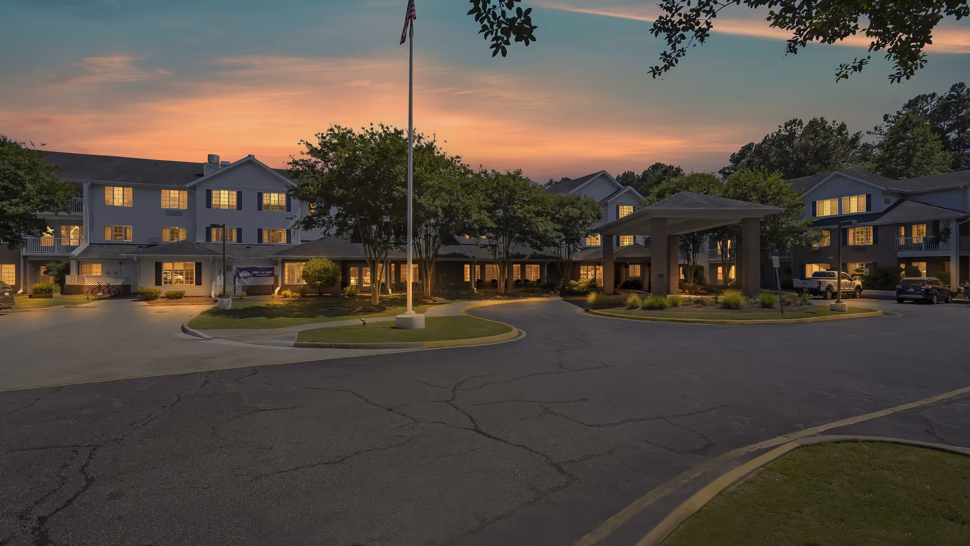 Exterior view of Lighthouse Pointe by Barclay House senior living facility at dusk, showing a large multi-story building with many lit windows, a covered entrance, trees, a flagpole, and a parking area with several cars.