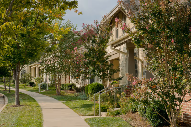 A sidewalk lined with trees and bushes runs alongside a row of residential buildings with front porches and stairs, surrounded by greenery and flowering plants under a partly cloudy sky.