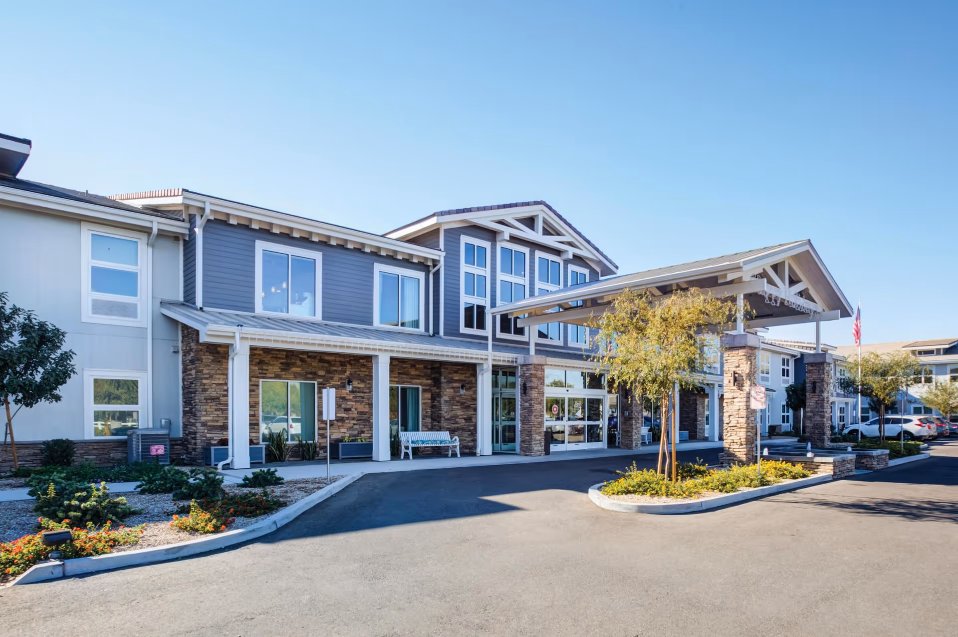 Front exterior of a modern two-story senior living building with a covered entrance, driveway and landscaping.