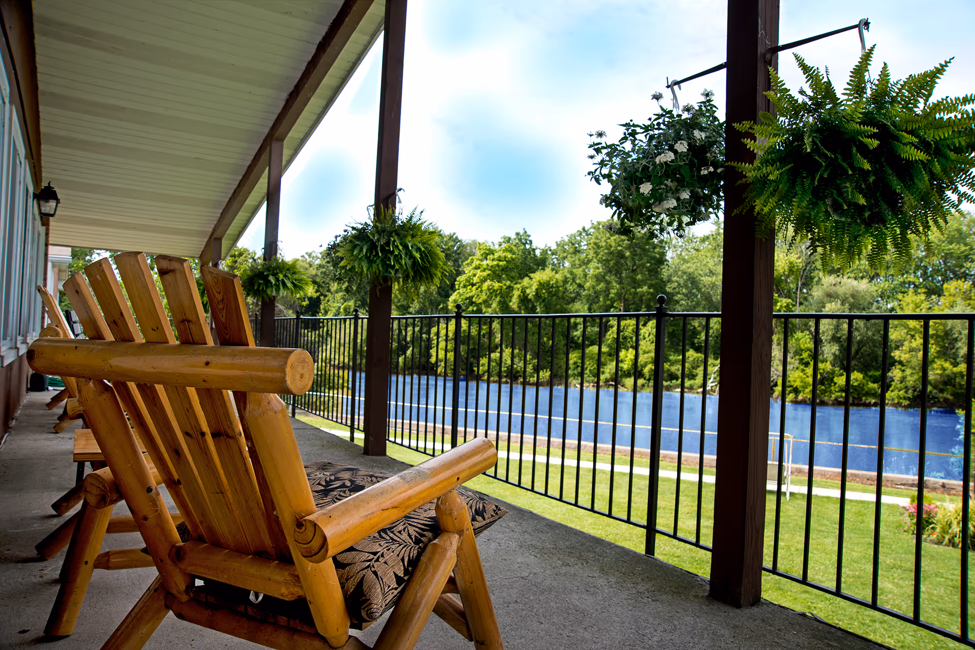 Wooden rocking chairs on a covered porch overlooking a river with green trees and hanging plants.