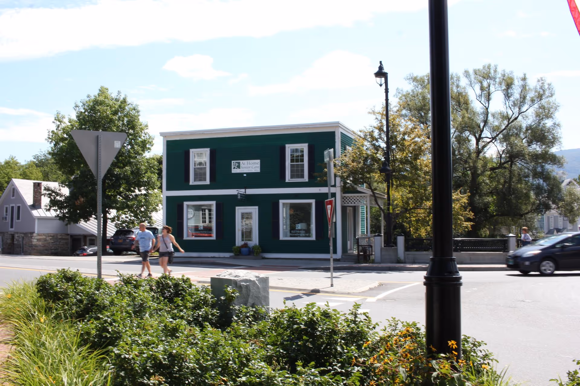 Green two-story storefront labeled "At Home Senior Care" on a street corner with pedestrians, a car, and landscaped foreground.