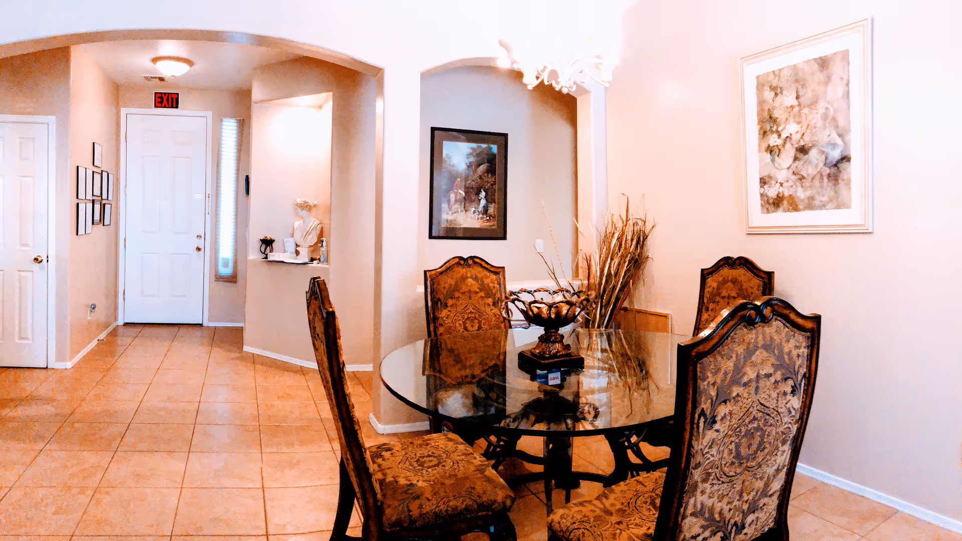 Interior view of a dining area with a round glass table surrounded by four ornate upholstered chairs. The room has beige walls with framed artwork, a decorative centerpiece on the table, and a tiled floor. An archway leads to a hallway with a white door and an exit sign above it.